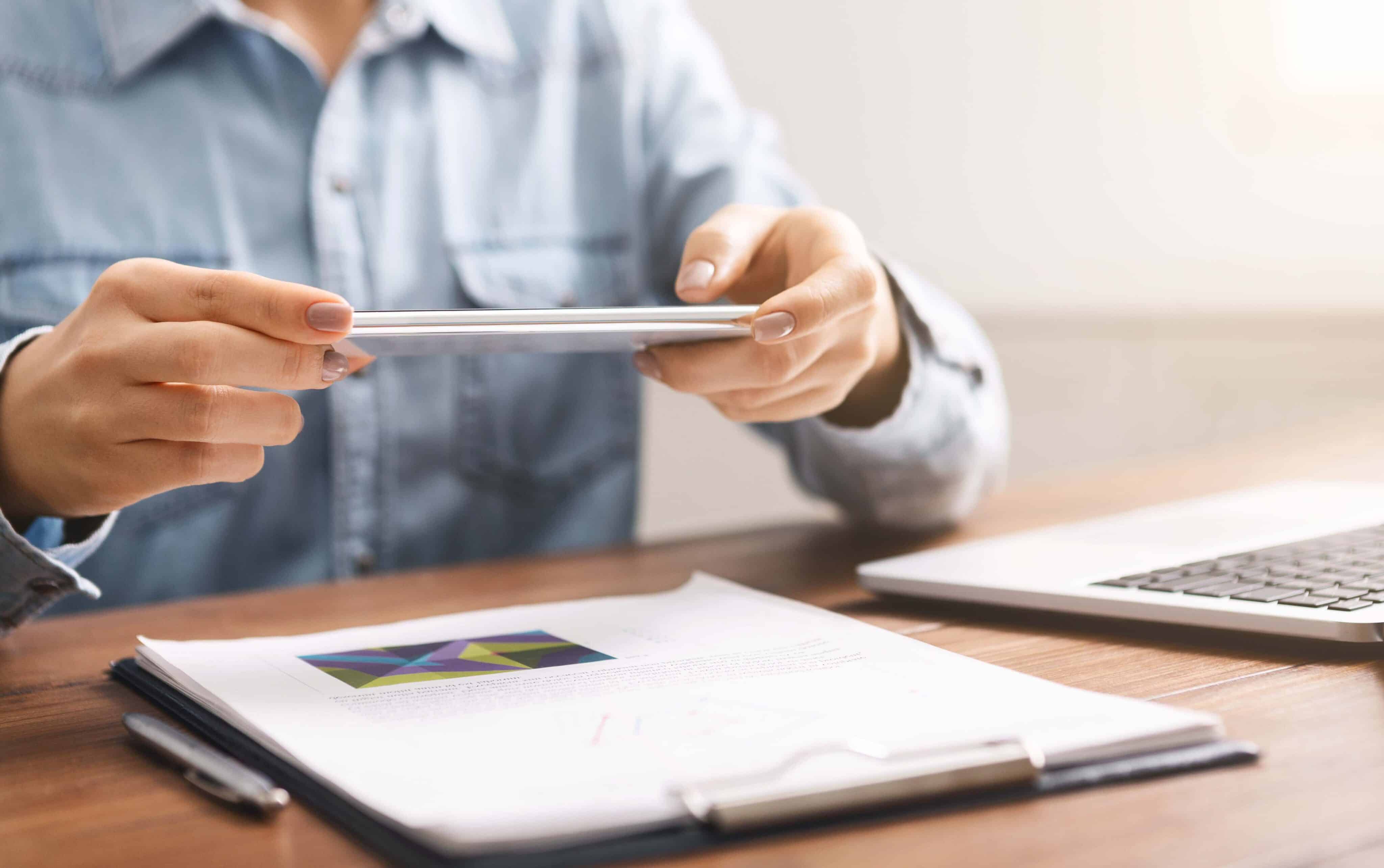 Mujer sosteniendo un teléfono inteligente horizontalmente tomando fotografías de documentos, usando una aplicación de escáner para hacer copias de documentos